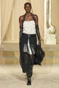 Model walks a runway in a strapless white beaded top with a black fringe skirt and long black opera gloves, boots visible underneath.