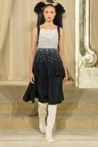 Model walking a runway in a black and silver sequin sleeveless dress with white knee-high boots on a stage backdrop.