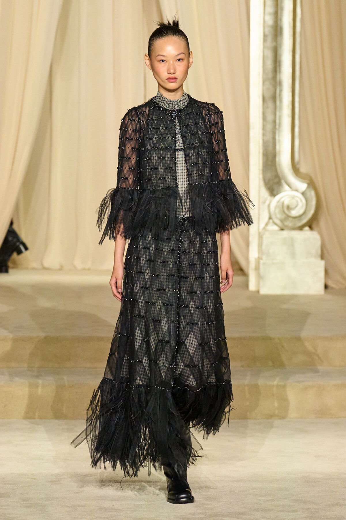 Model walking a runway in a black, floor-length dress with sheer polka-dot sleeves, beaded panels, and feathered hem; heavy boots visible.