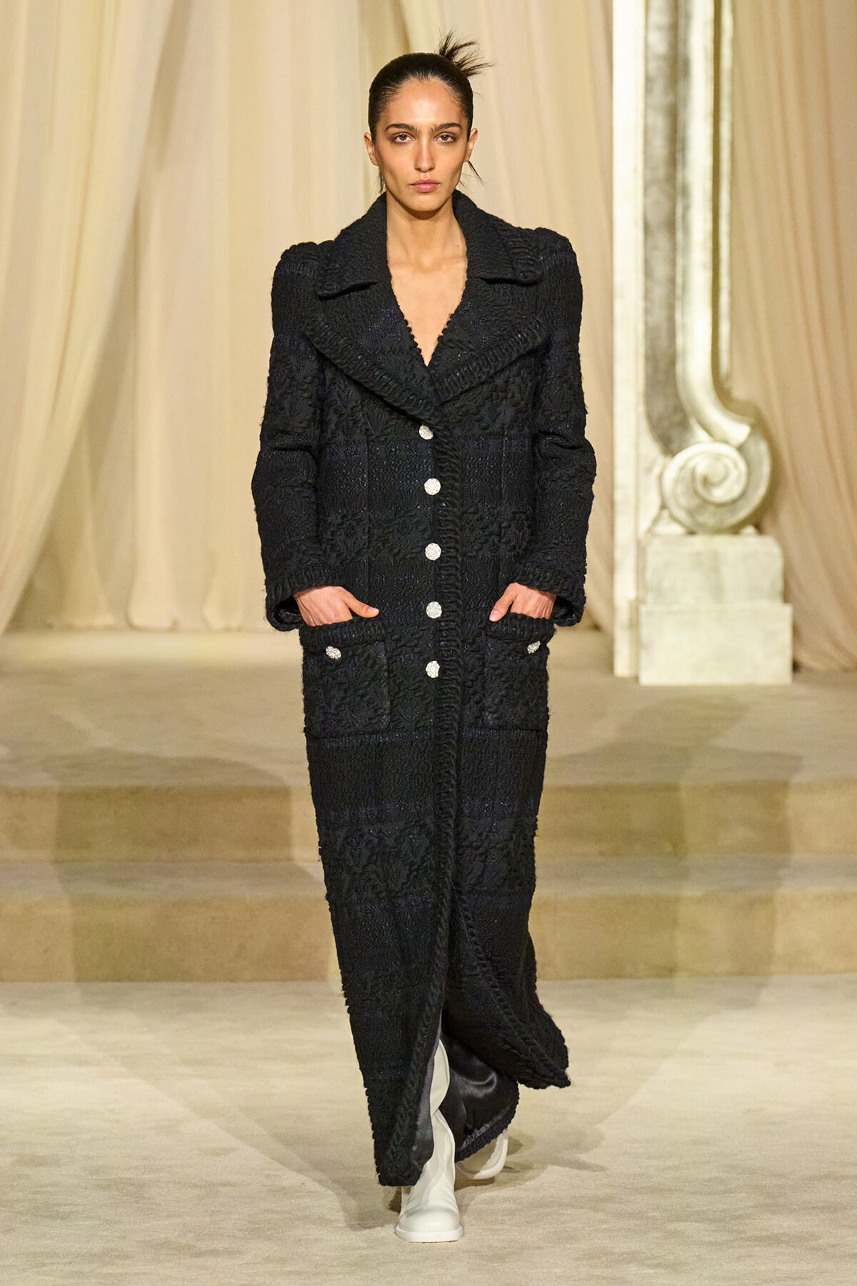 Model walking the runway in a full-length black textured coat with oversized lapels, white buttons, and pockets, paired with white boots against a draped backdrop.