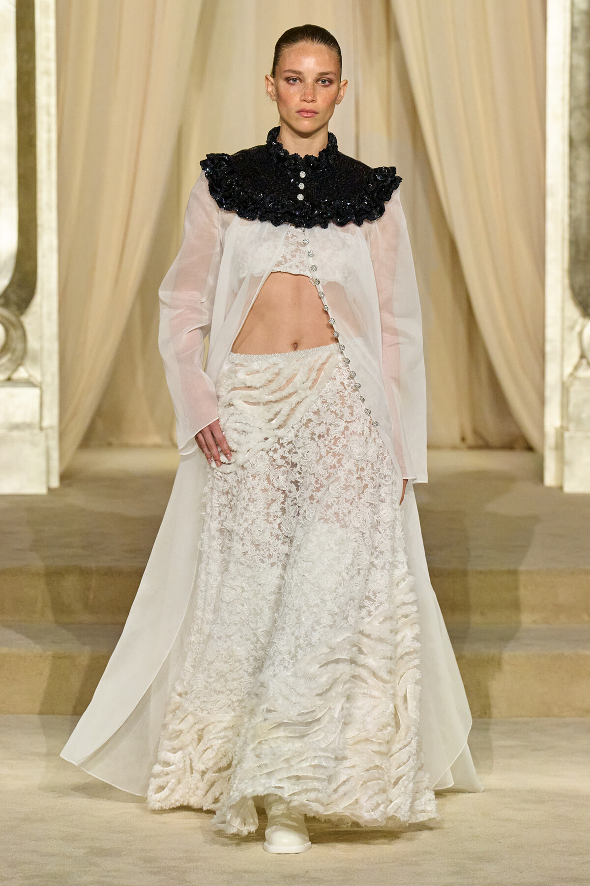 Model walking a runway in a sheer white blouse with a dramatic black ruffled collar and an off-white lace skirt with a diagonal beaded detail.