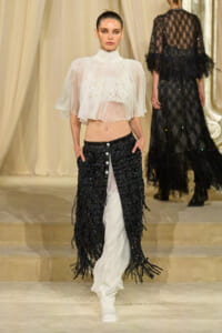 Model walks a runway in a sheer white cropped blouse with ruffled cape sleeves and black sequin fringe pants, beige backdrop behind.