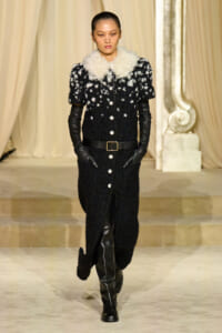 Model walks a runway in a black textured coat with a white fur collar, belted waist, black gloves, and knee-high boots.