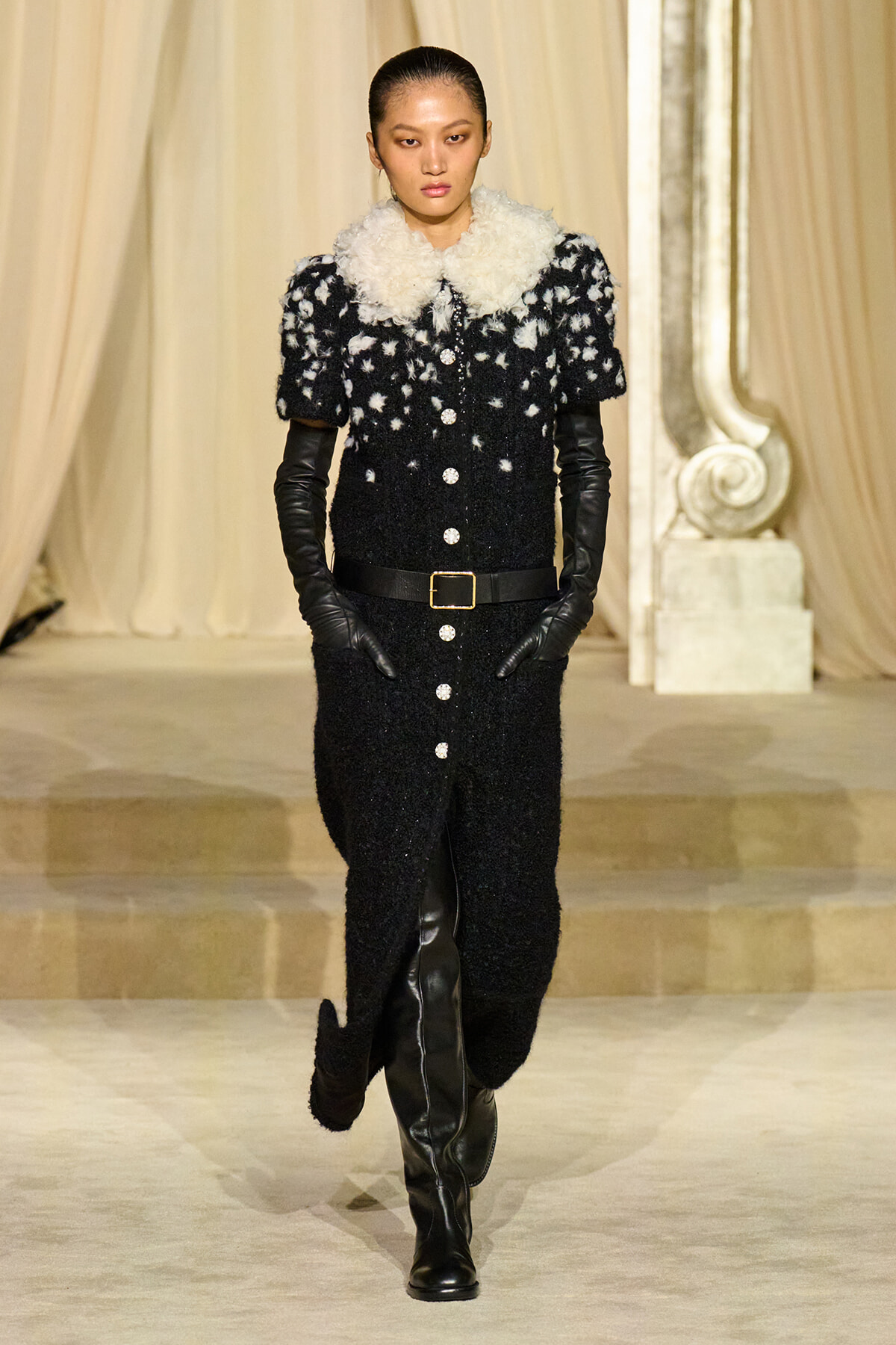 Model walks a runway in a black textured coat with a white fur collar, belted waist, black gloves, and knee-high boots.