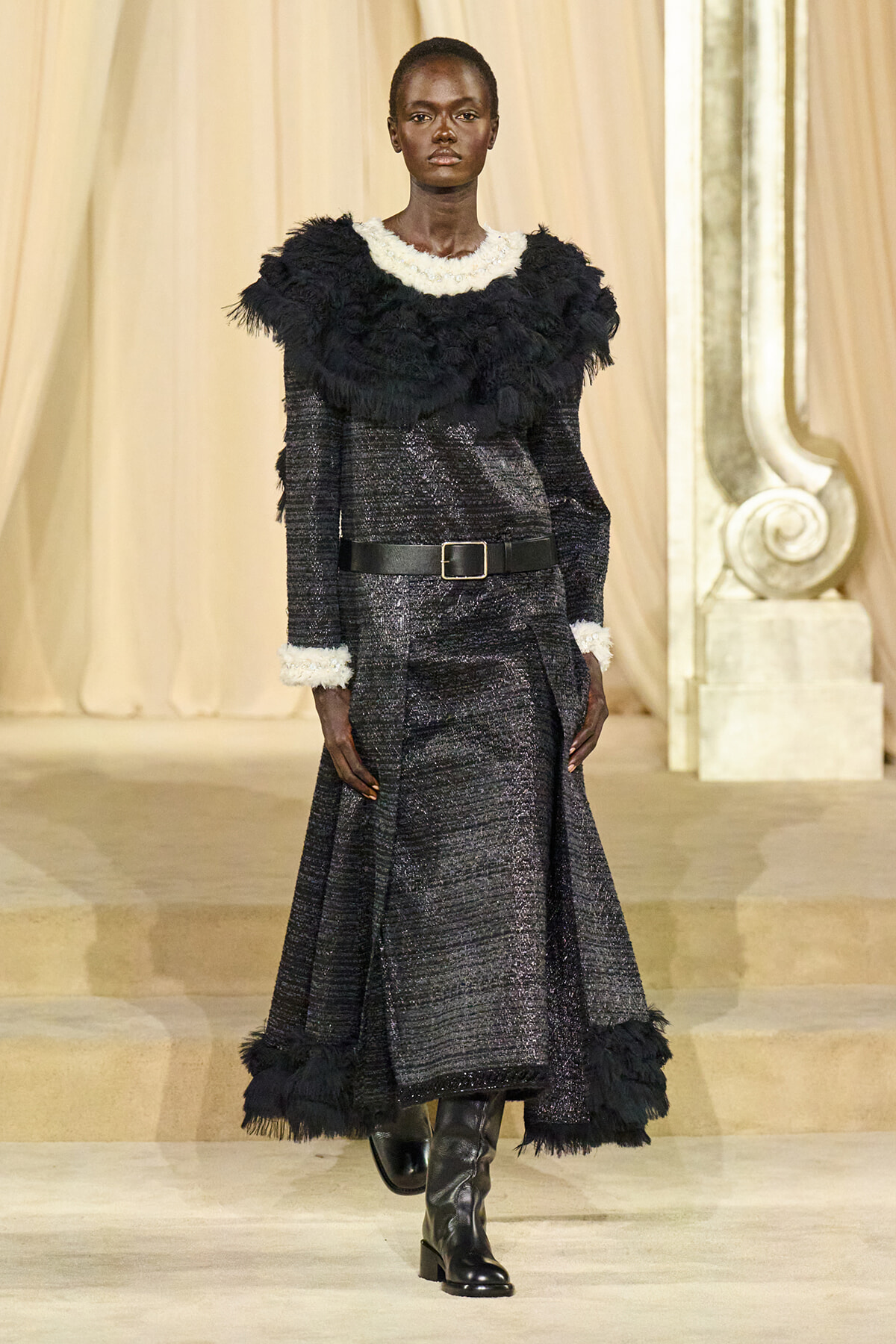 Model walks the runway in a long black textured gown with white fur trim at the neckline and cuffs, cinched with a wide belt and knee-high boots.