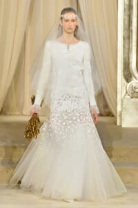 Bride-model in a white beaded wedding gown with long sleeves and a sheer veil, holding a textured gold bouquet on a runway.