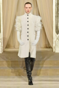 Model on runway wearing a long ivory textured coat with feathered sleeves, black buttons, white gloves, and black over-the-knee boots on a beige stage.