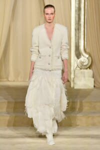 Model on a runway wearing a white textured blazer with silver buttons and a multi-tiered feathered white skirt.