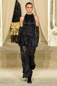 Model walks a runway in a black satin halter top with ruched bodice and layered details, paired with sheer lace pants and long black gloves.