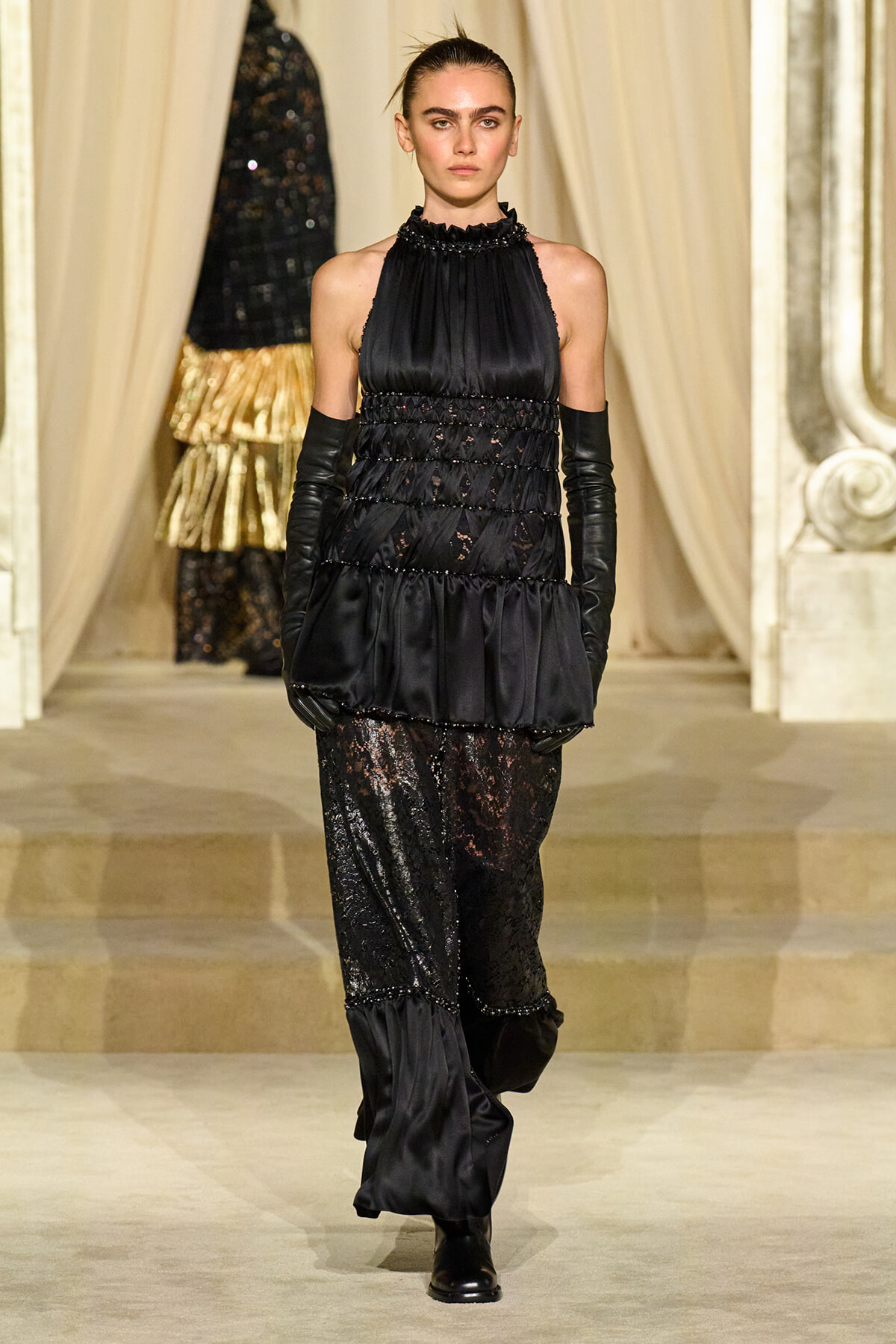 Model walks a runway in a black satin halter top with ruched bodice and layered details, paired with sheer lace pants and long black gloves.