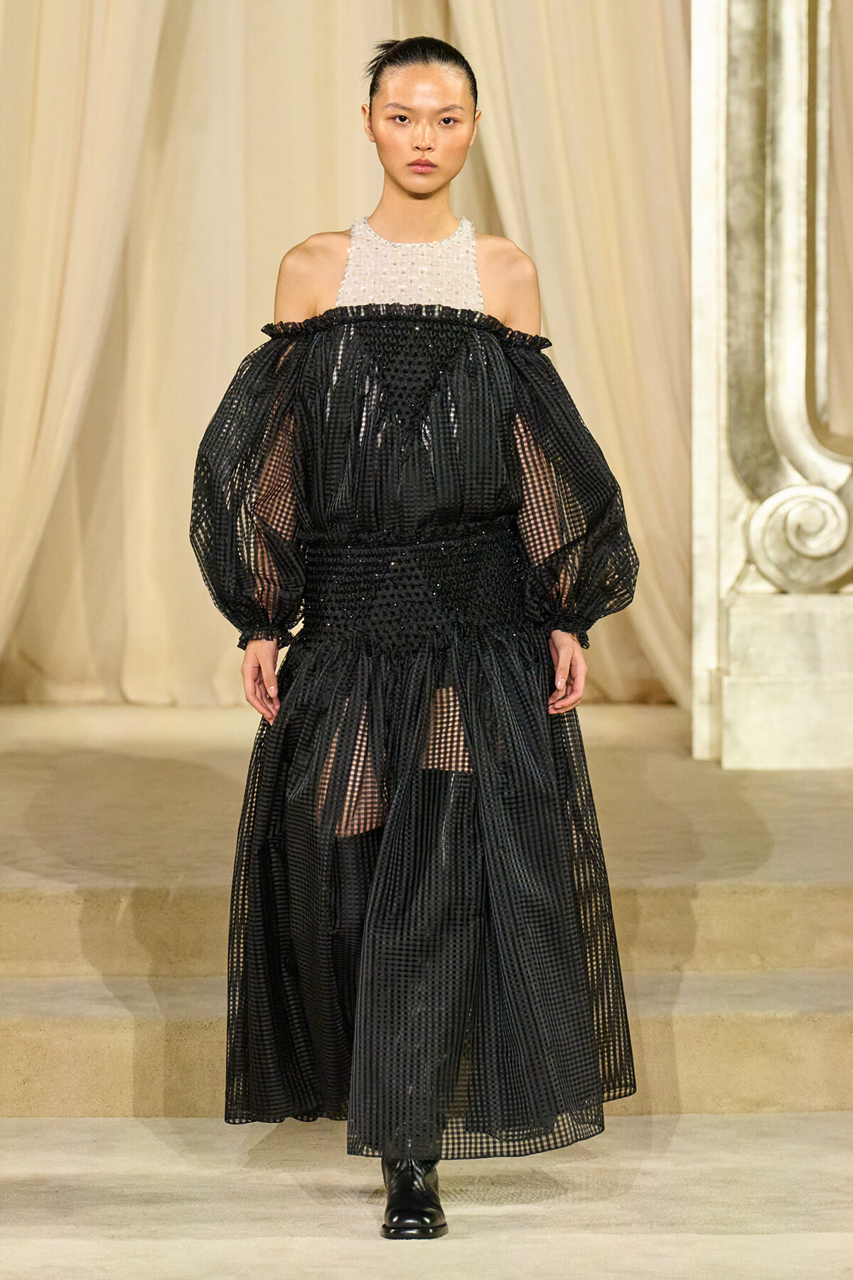 Runway model wearing a sheer black dotted gown with off-the-shoulder neckline and white beaded halter top, voluminous sleeves.