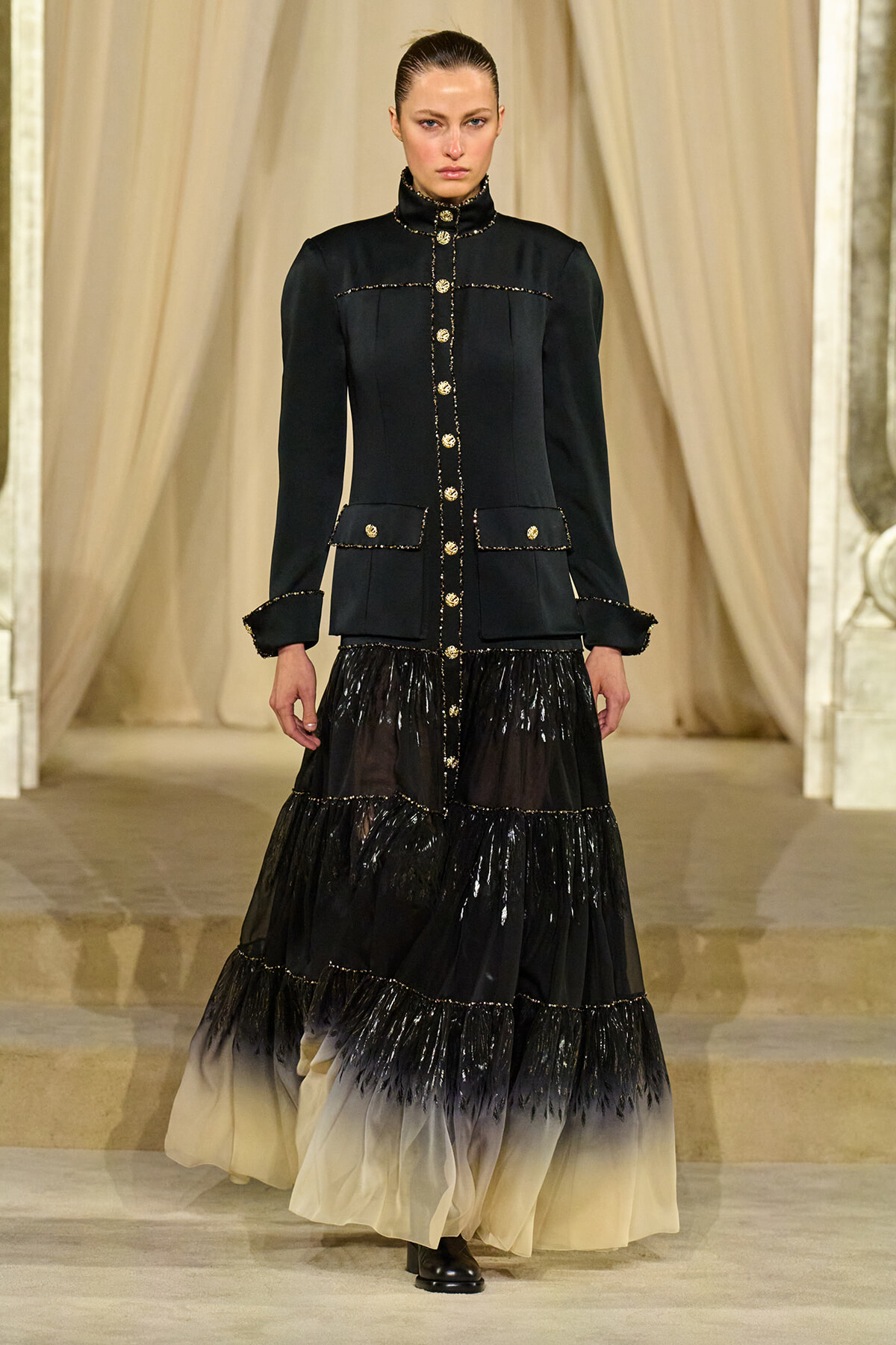 Model on runway wearing a black military-style coat with gold buttons and a tiered gradient skirt fading from black to cream.