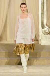 Model walking a runway in a sheer ivory dress with a quilted bodice, gold metallic ruffled hem, and white knee-high boots on a neutral stage.