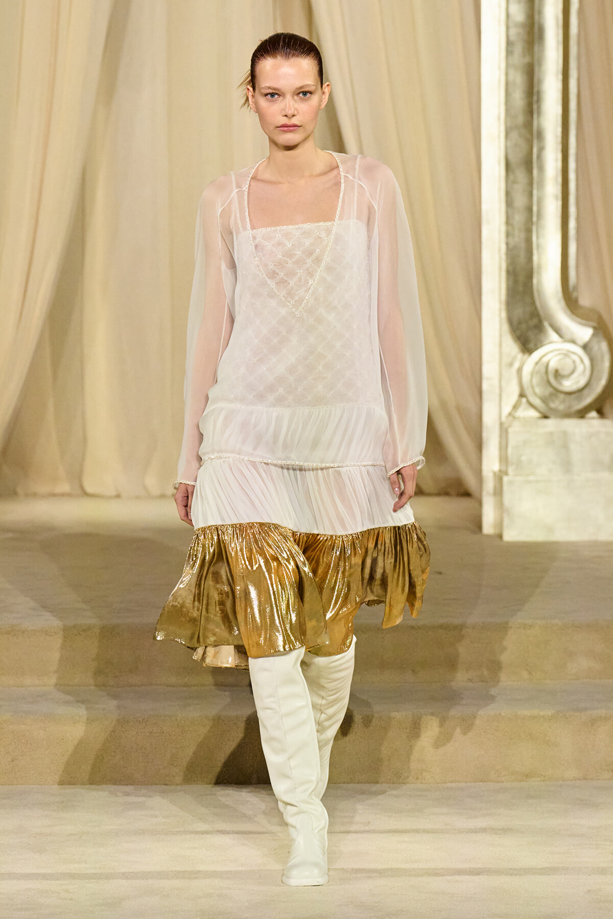 Model walking a runway in a sheer ivory dress with a quilted bodice, gold metallic ruffled hem, and white knee-high boots on a neutral stage.