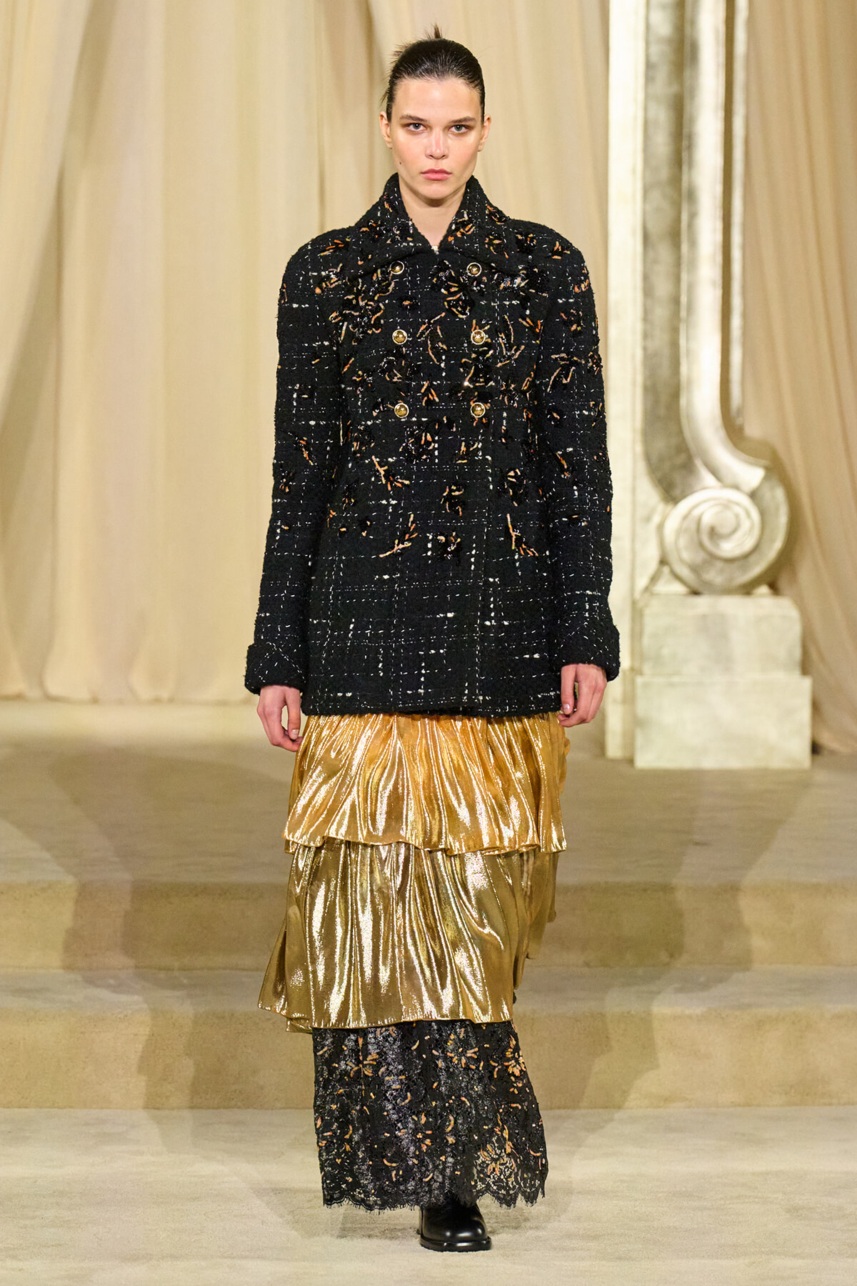 Runway model in a black textured coat with gold flecks and a gold layered metallic skirt on a fashion show catwalk.