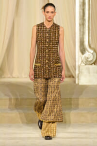 Model walks the runway in a sleeveless brown-gold tweed top and matching wide-leg tweed pants, beige draped backdrop in the background.