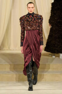 Model walking a runway in a burgundy sequin-embellished top with a draped satin skirt and tall black boots.