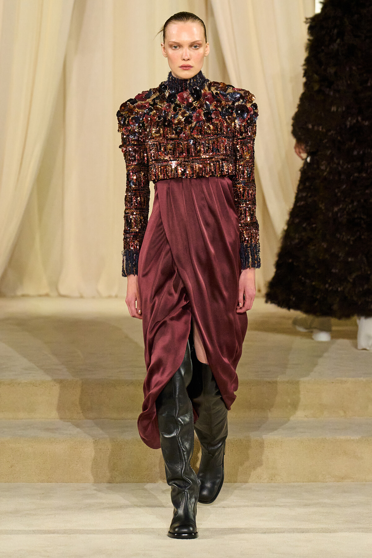 Model walking a runway in a burgundy sequin-embellished top with a draped satin skirt and tall black boots.