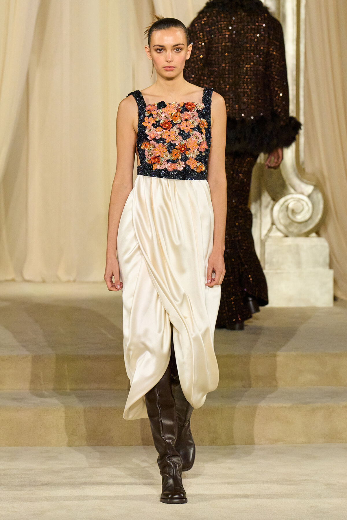 Model walking the runway in a cream satin draped skirt with a navy floral-embellished bodice and knee-high brown boots.