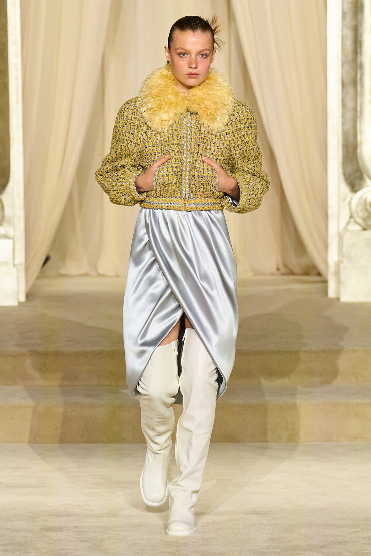 Model walking a runway in a yellow textured jacket with a fluffy fur collar, a silver draped skirt, and white knee-high boots.