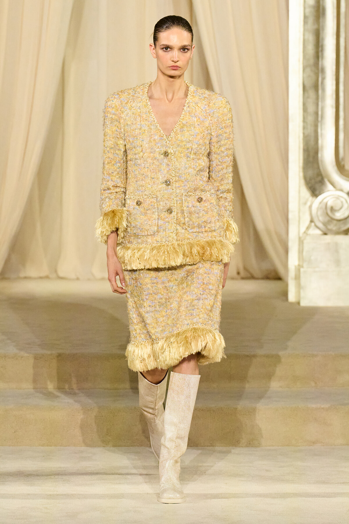 Model on runway wearing a cream tweed suit with fringe trim at cuffs and hem, paired with beige knee-high boots.