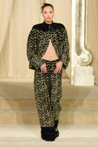 Model on a runway wearing a black-and-gold patterned cape with an open front revealing midriff and matching wide-leg pants, black fringe boots.