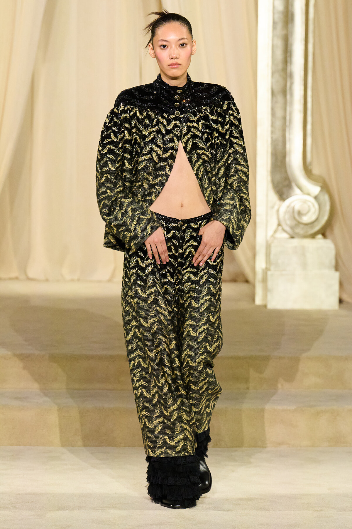 Model on a runway wearing a black-and-gold patterned cape with an open front revealing midriff and matching wide-leg pants, black fringe boots.