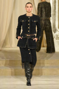 Model walking a fashion runway in a black tweed suit with gold buttons and braided trim, paired with a black belt and knee-high boots.