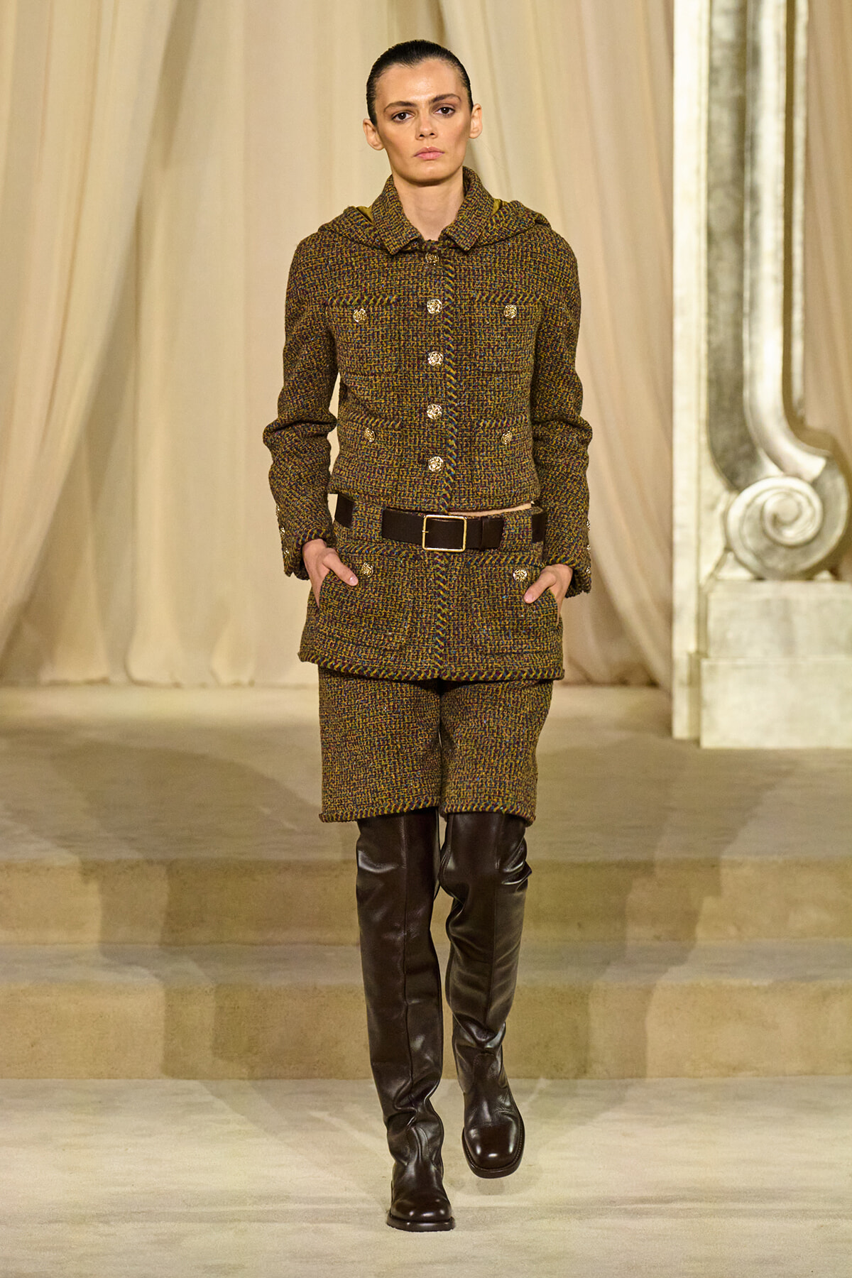 Model walking the runway in a brown tweed jacket and shorts set with a wide black belt and tall dark boots, posing on a fashion show runway of draped backdrop.