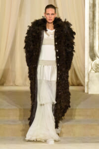 Model on a runway wearing a white tiered gown with silver beading and a long black feathered coat.