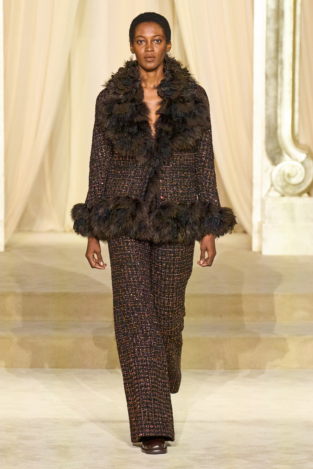 Model walks the runway in a dark tweed suit with heavy black fur trim at the collar, cuffs, and hem.