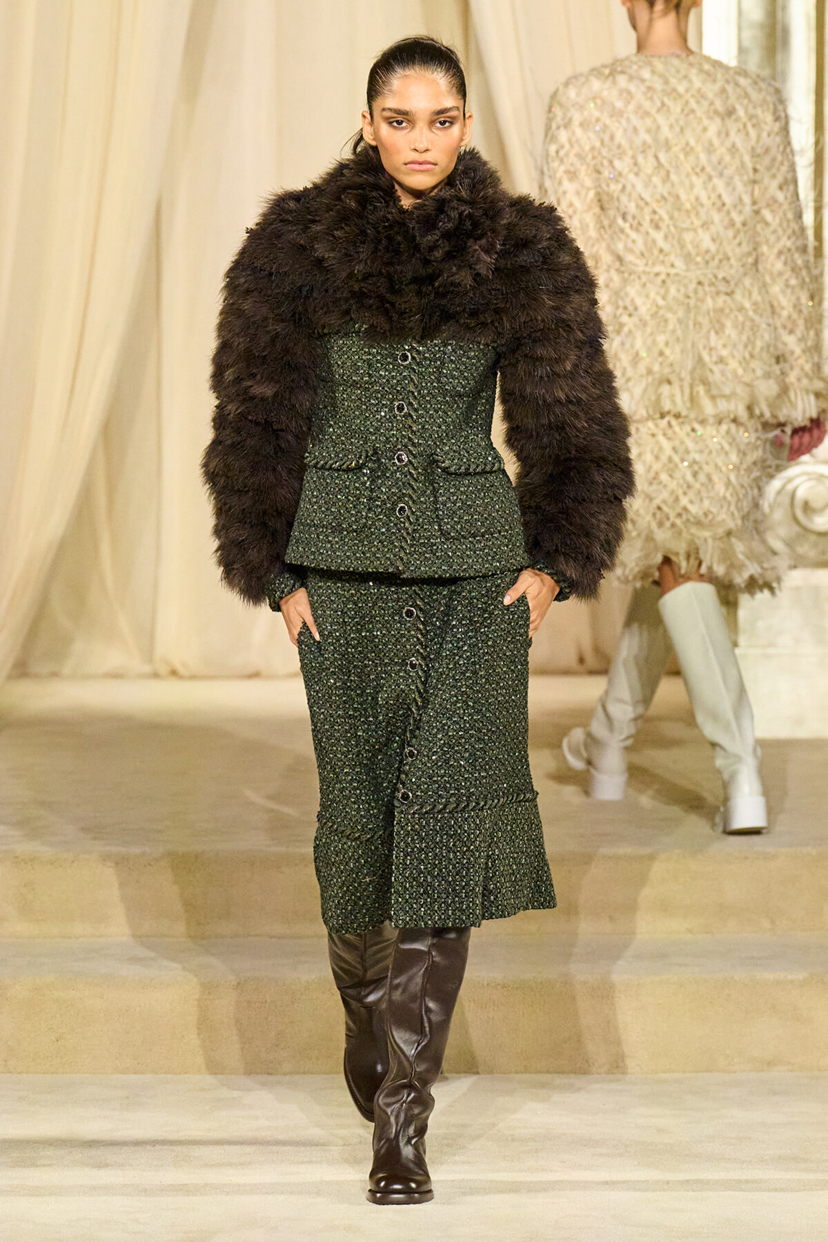 Model walks runway wearing a dark green textured suit with a large black fur stole and tall brown boots.