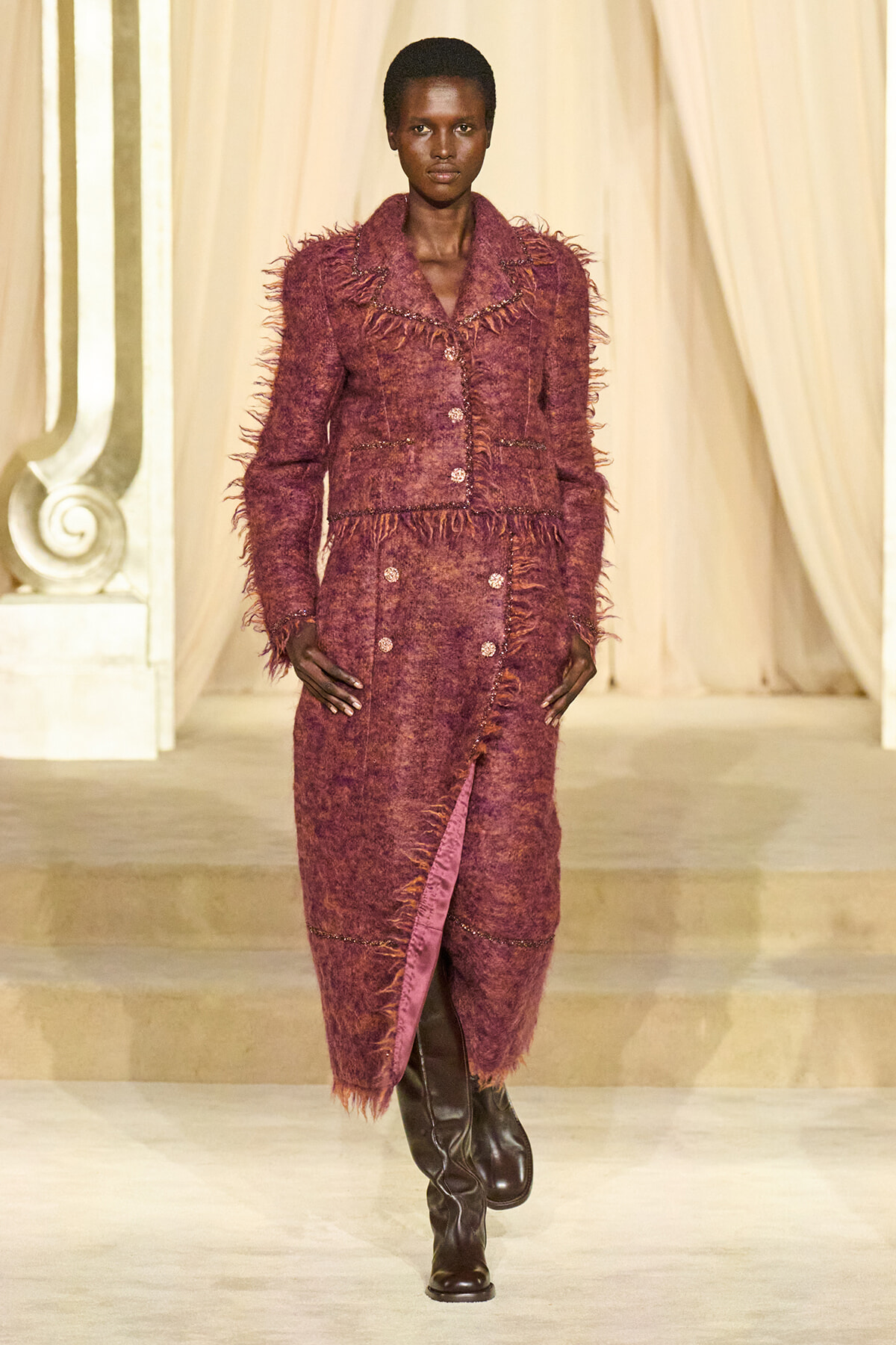 Model walks a fashion runway wearing a burgundy fringed textured coat with matching dress and knee-high black boots.