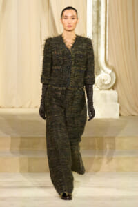 Model on a runway wearing a dark green textured tweed suit with frayed edges and long black gloves, walking forward confidently.