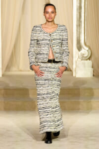 Model walking a fashion runway in a cream and black tweed two-piece suit with a cropped jacket, high-waisted long skirt, black belt, and ankle boots.