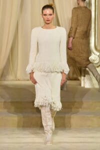 Model walks the runway in a white textured knit dress with fringe at the cuffs and hem, paired with knee-high boots.