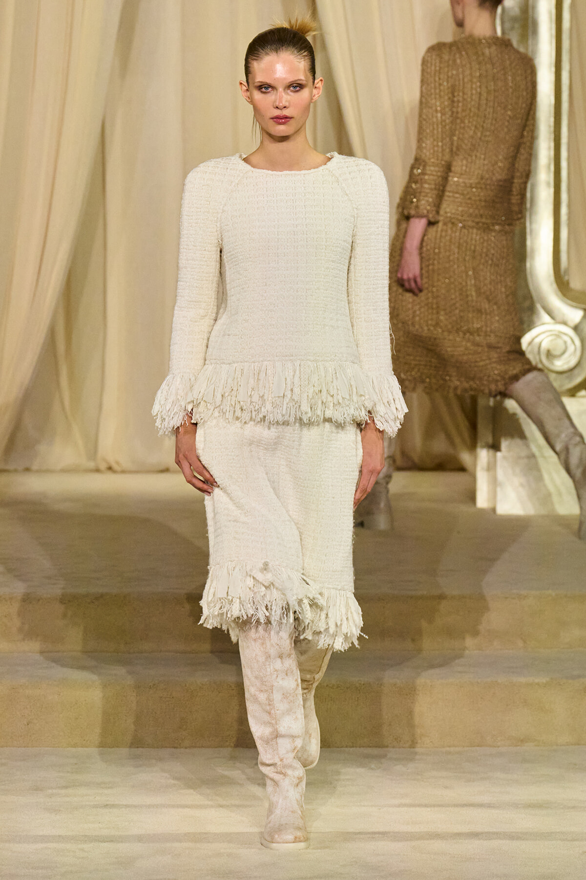 Model walks the runway in a white textured knit dress with fringe at the cuffs and hem, paired with knee-high boots.