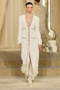 Model walks a runway wearing a cream textured long coat with gold trim and fringe at the hem, paired with light boots.