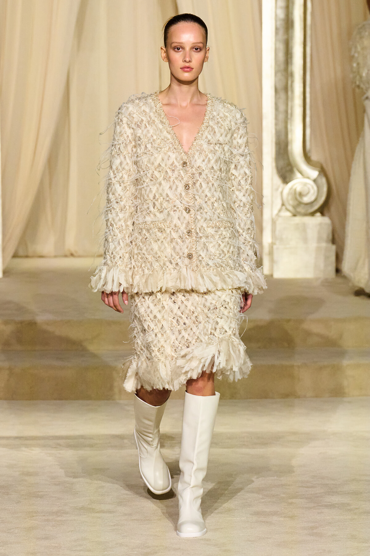 Model walking a fashion runway in a cream boucle suit with fringe details and a matching skirt, paired with white knee-high boots