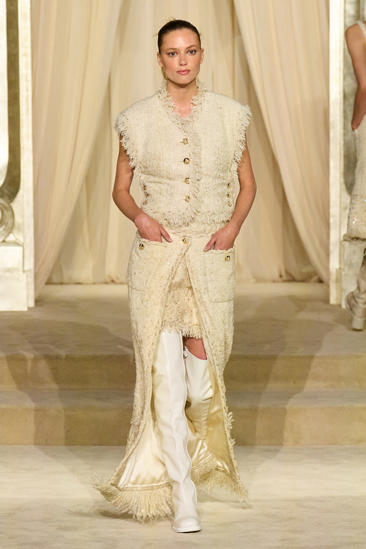 Model walks a fashion runway in an ivory textured fringe suit with gold buttons and knee-high white boots, hands in pockets.
