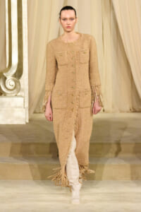 Model on a runway wearing a tan tweed long coat with gold buttons, fringe cuffs, and fringe hem over knee-high boots.