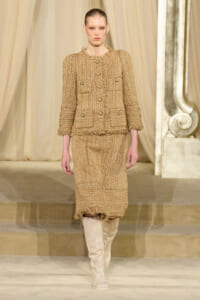 Model walks a runway in a tan tweed fringe-trimmed suit with gold buttons, paired with knee-high beige boots.