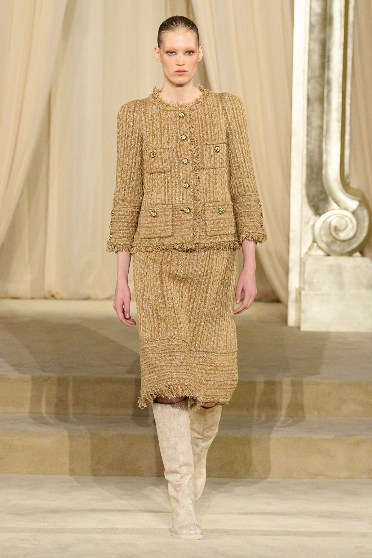 Model walks a runway in a tan tweed fringe-trimmed suit with gold buttons, paired with knee-high beige boots.