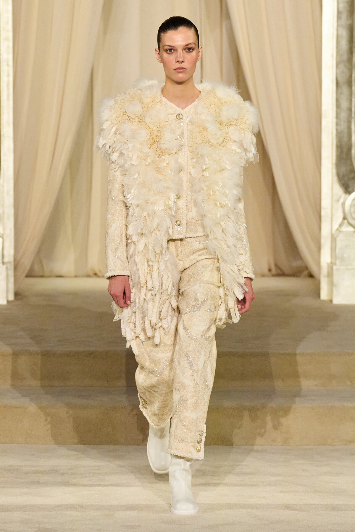 Model walks the runway in an ivory feathered coat over a cream embellished suit, with white boots.