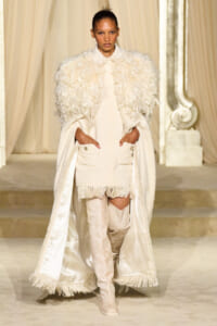 Model walking a runway in an all-white textured ensemble with feathered cape, fringed hem dress, and over-the-knee boots in a cream setting.