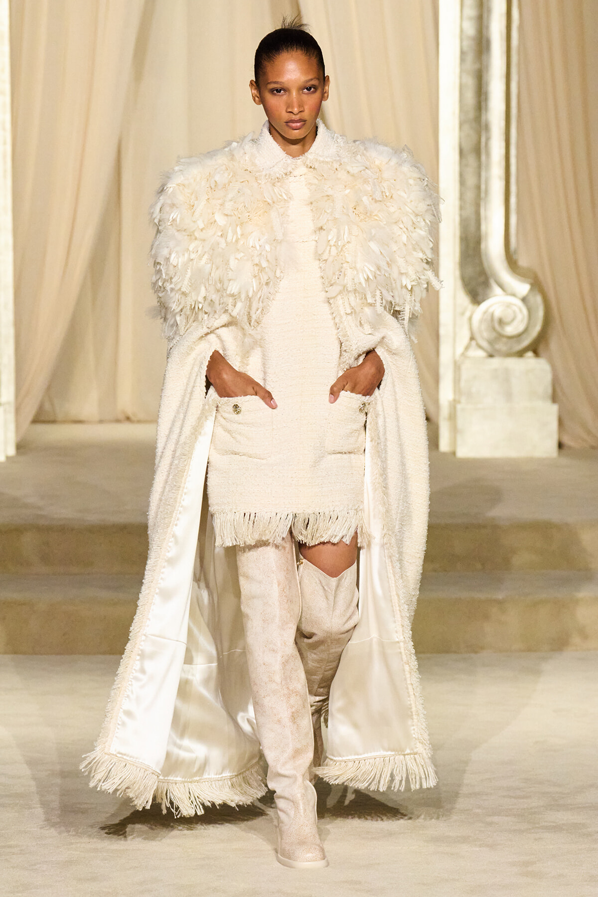 Model walking a runway in an all-white textured ensemble with feathered cape, fringed hem dress, and over-the-knee boots in a cream setting.