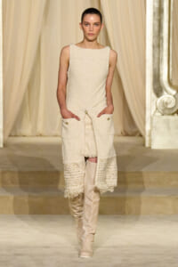 Female model walking a cream textured sleeveless dress with a long, fringed coat and matching boots on a runway.