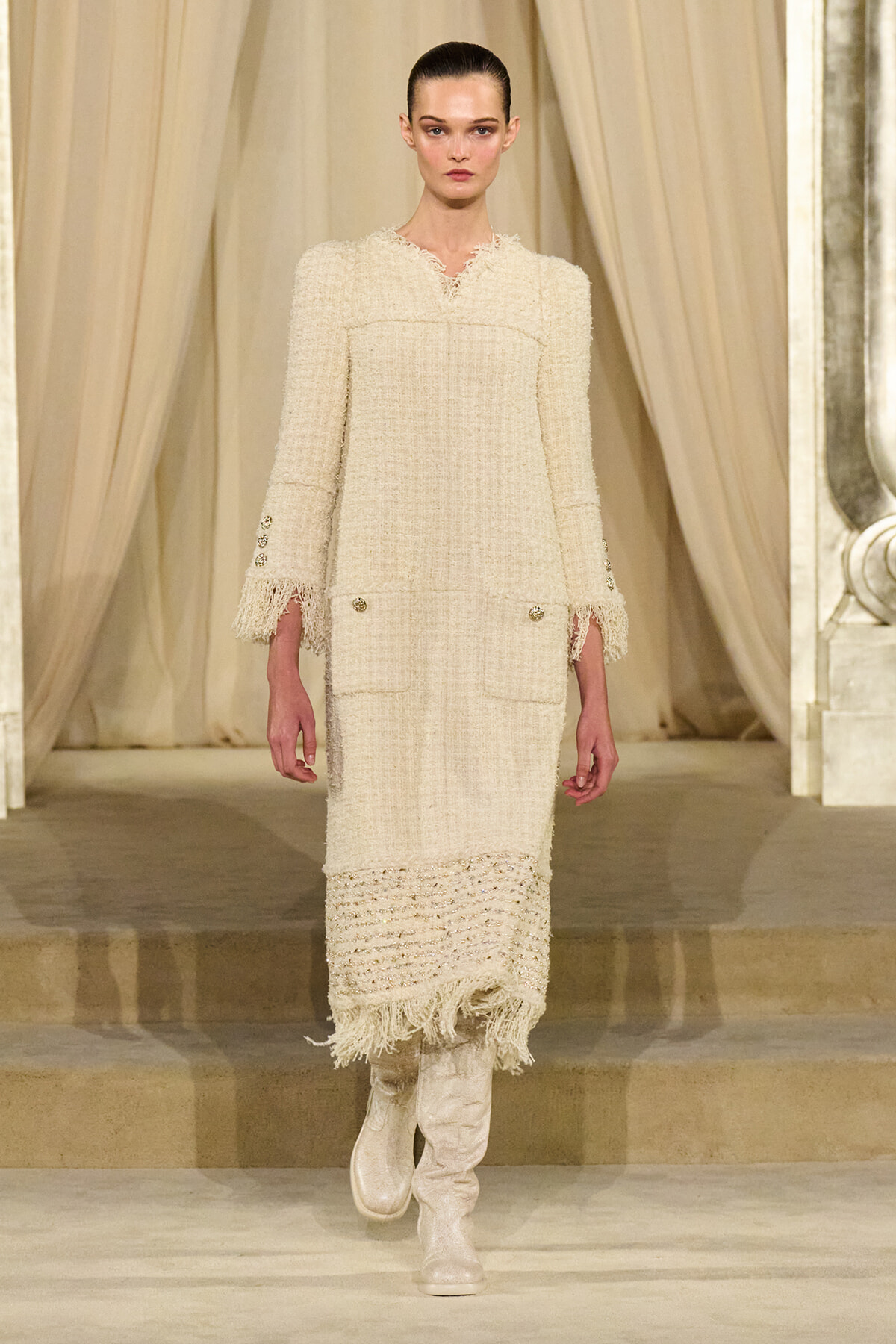 Model walking a runway in a cream textured boucle coat dress with fringe cuffs and hem, paired with white knee-high boots.