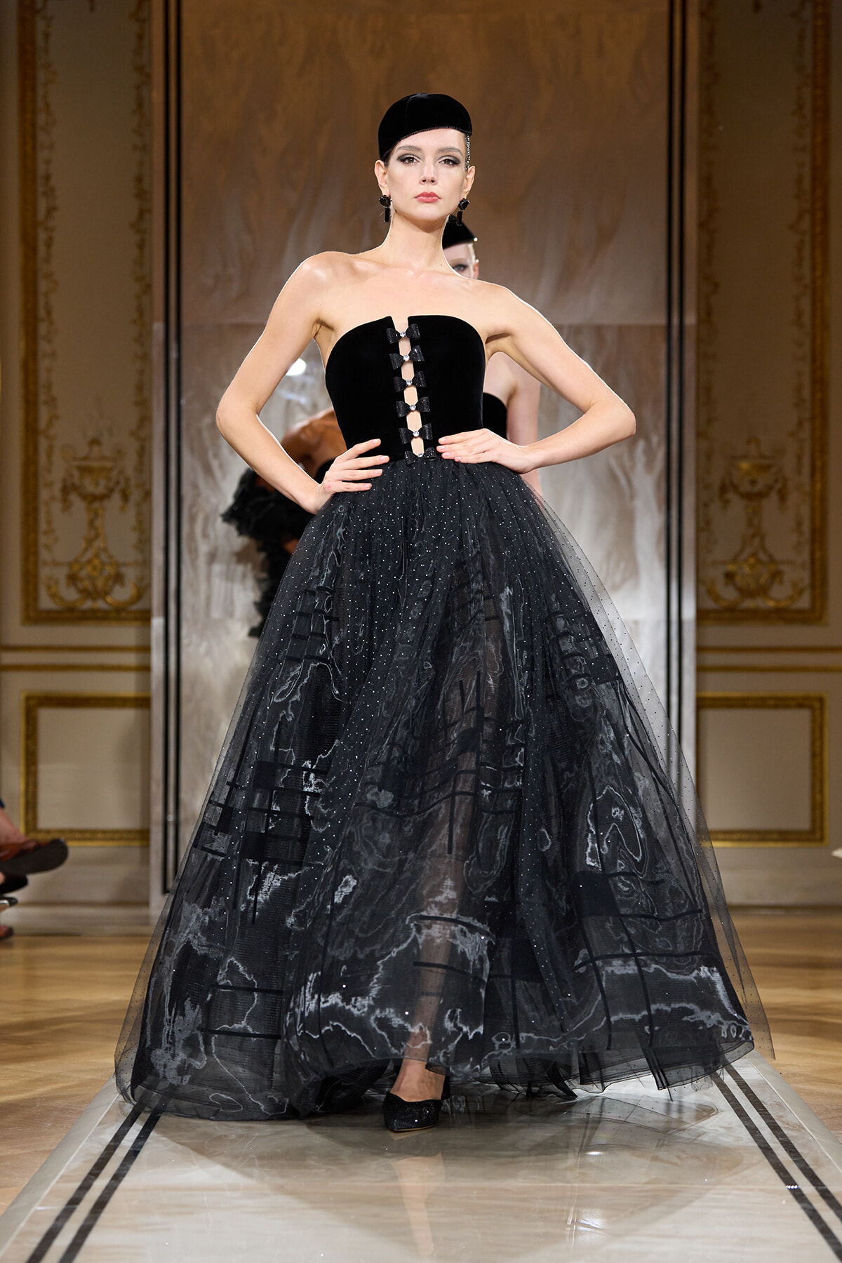 Model on runway wearing a strapless black ball gown with a jeweled front corset and voluminous tulle skirt.