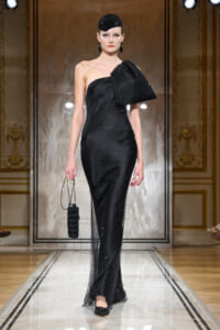 Model walks a runway in a black one-shoulder sequined gown with a large bow at the shoulder, paired with a black cap and matching clutch.